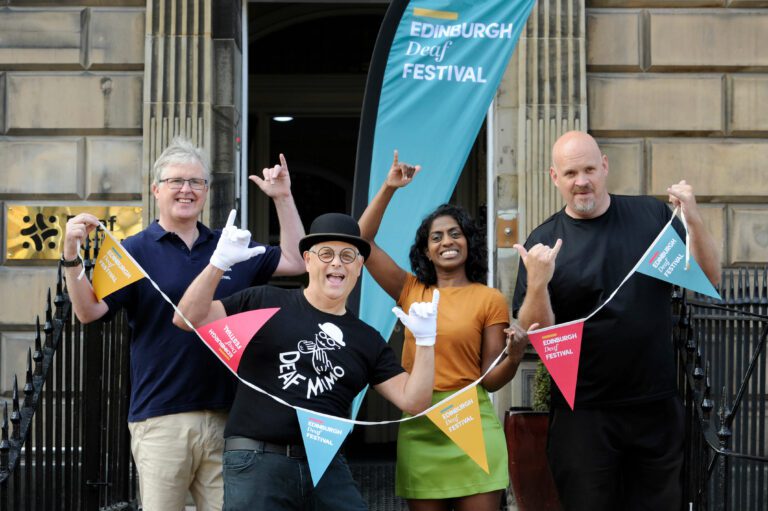 People standing on the steps of the Deaf Action offices, holding bunting and standing in front of a banner for Edinburgh Deaf Festival. One of the people is wearing a black bowler hat, white gloves and a t shirt which says Deaf Mimo.