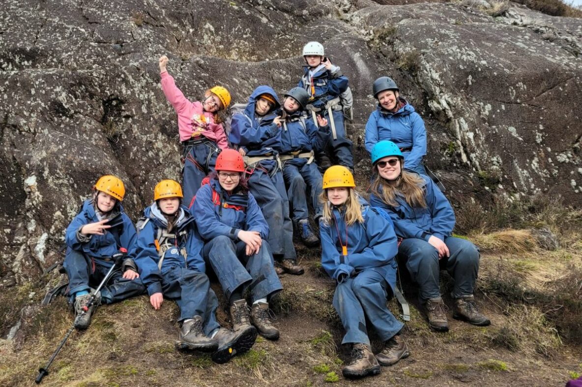 A group of young people stand beside a large rock. They are wearing outdoor clothing and climbing helmets.