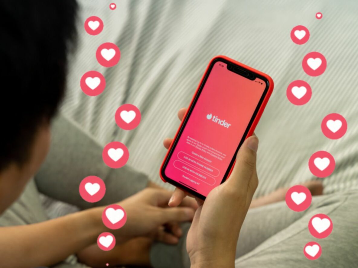 Person looking at phone showing the Tinder app homescreen - a red background with Tinder written across the centre of the screen, followed by terms and conditions text below this. Surrounding the phone is a series of white love hearts in red circles.