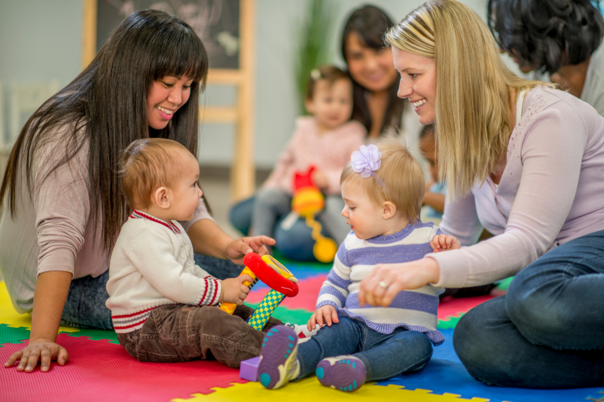 Parent and toddler group. Three females sat on a multicoloured soft play floor mat alongside young children while playing with colourful toys.