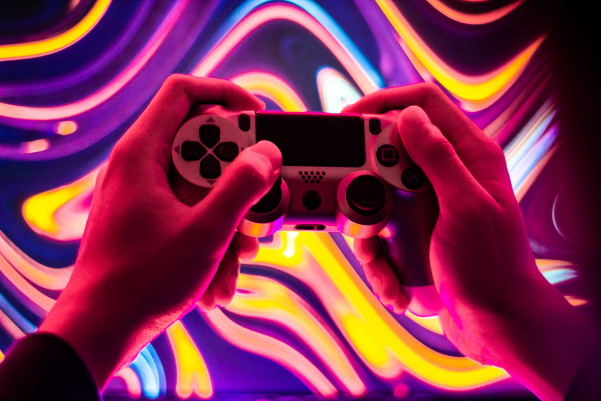 A video game console controller is held between two hands, behind is a colourful screen of swirling colours.