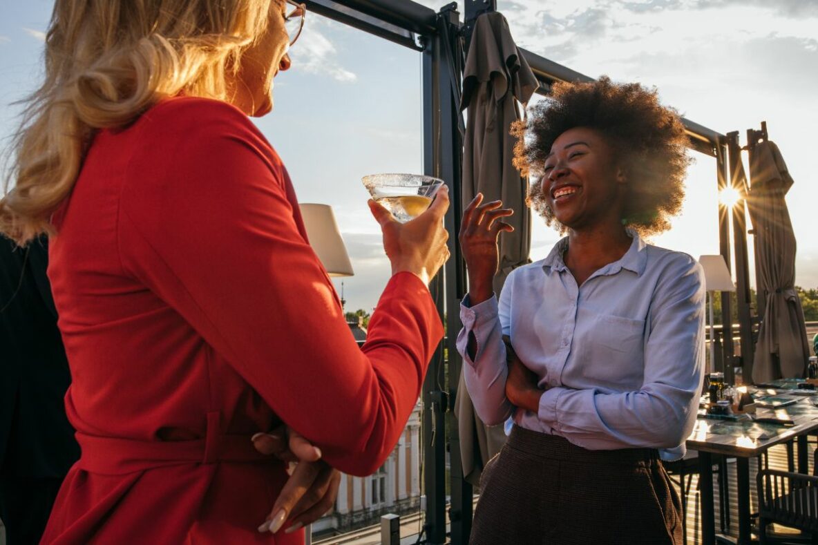 Two women stand on a roof top bar talking, one of them is holding a drink in their hand.