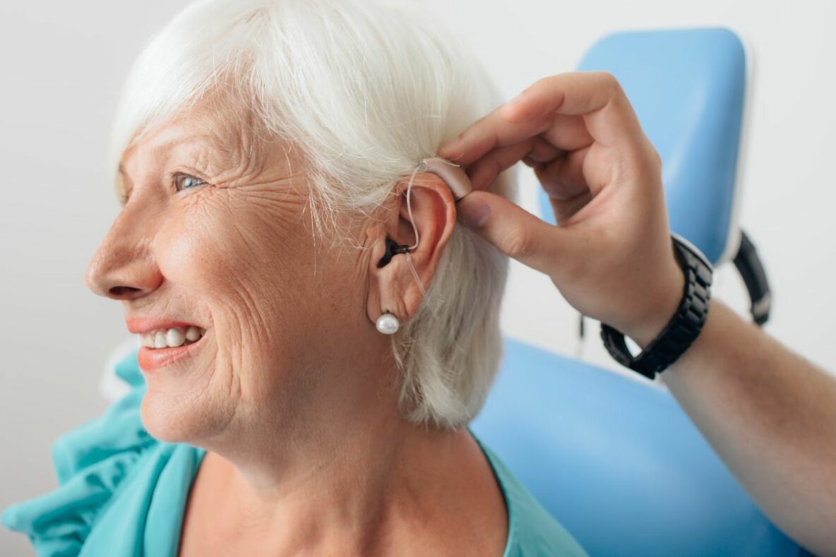 An older woman sits in a chair whilst another person adjusts her hearing aid.