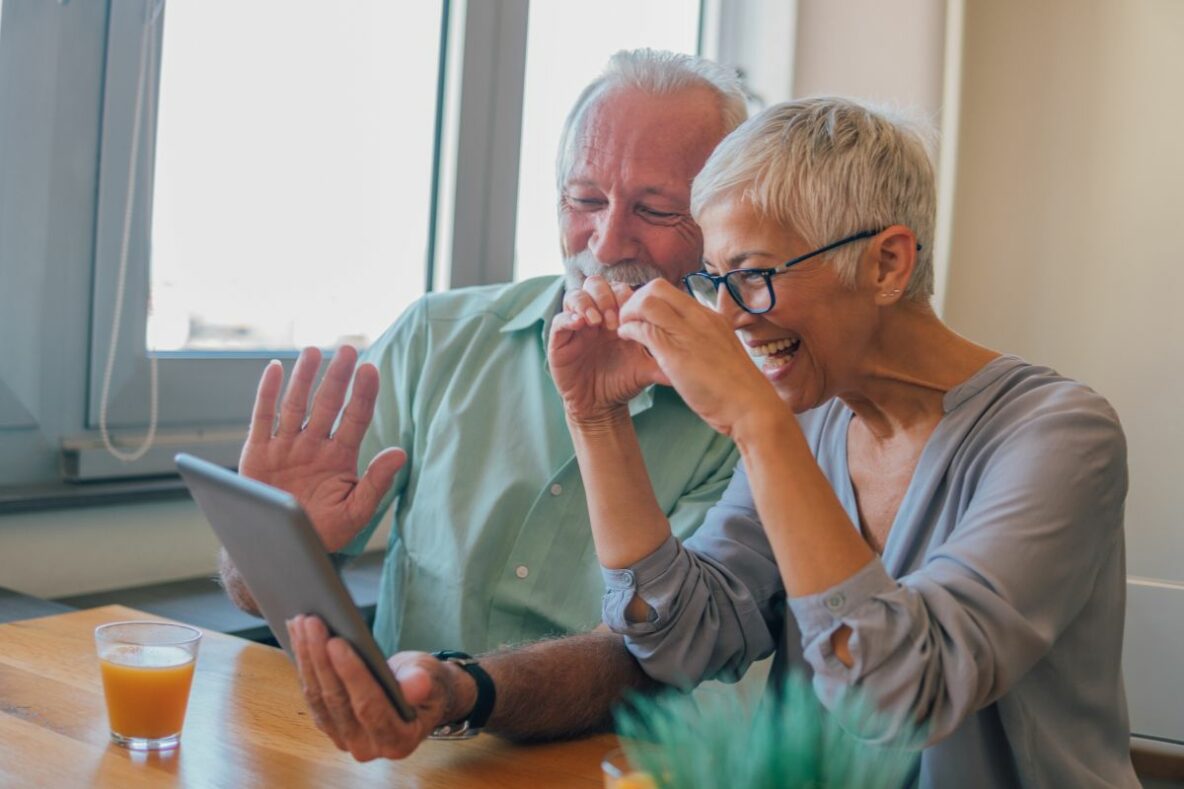 Two older adults sitting at a kitchen table. They are waving to someone on the other end of a video call that is taking place on a tablet.