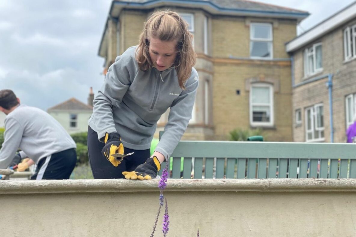 A woman with long blonde hair is wearing gardening gloves and repairing a damaged wall. Behind her someone else is gardening. There is a large house in the background and flowers in the foreground.