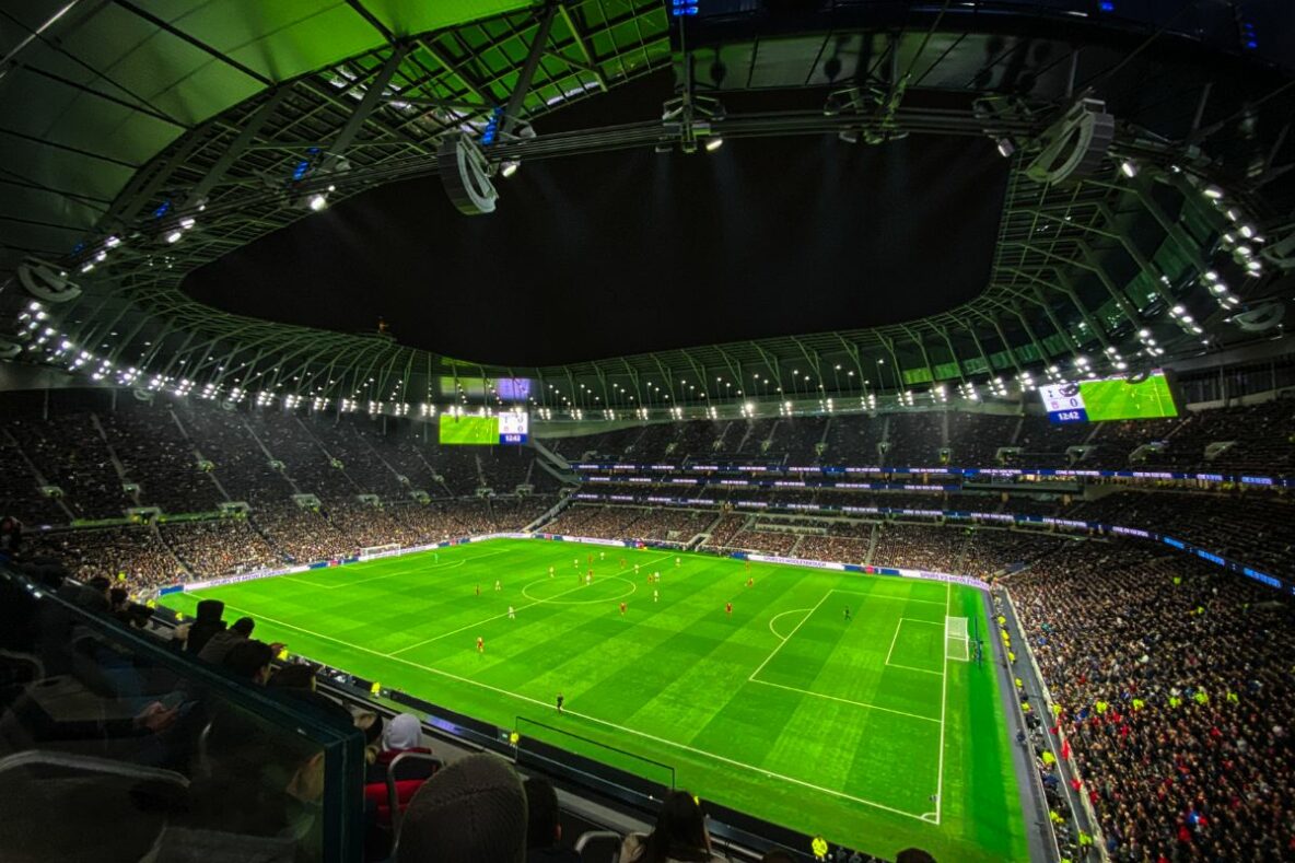 A football stadium at night, a huge number of supporters watch from the stands as the illuminated pitch has two teams playing against one another.