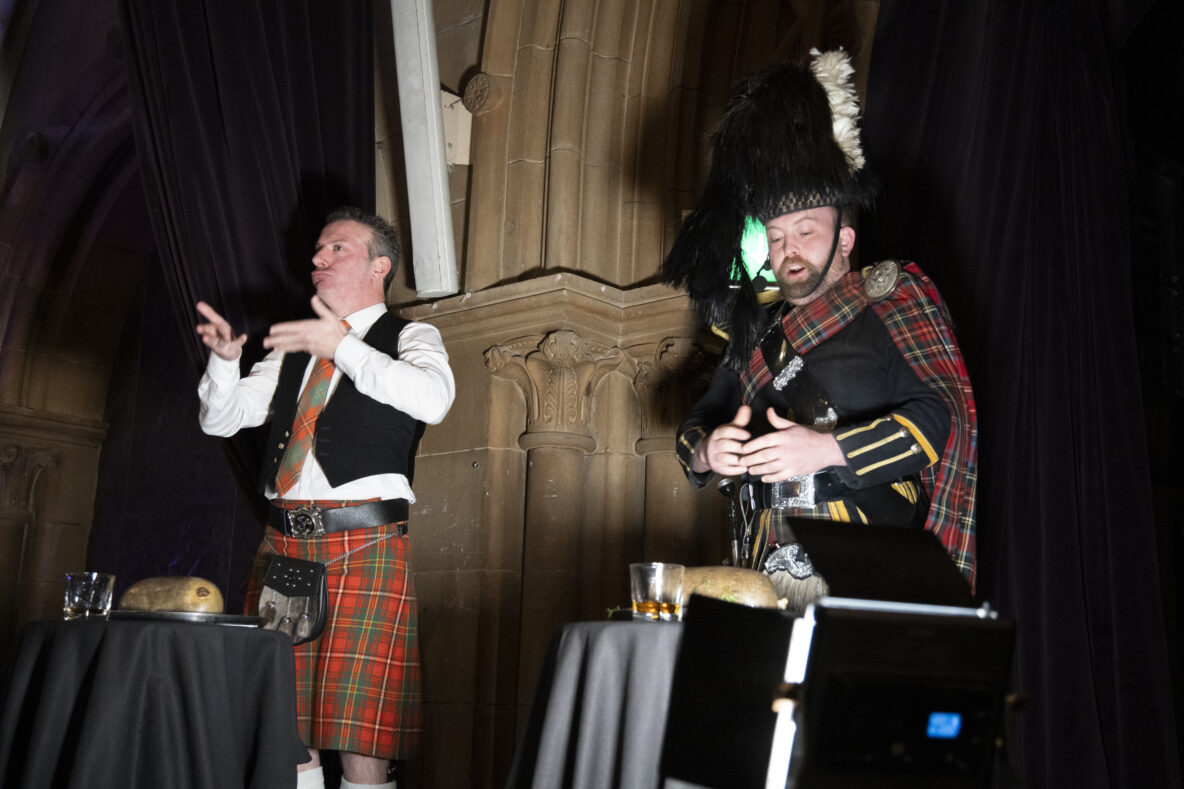 Man in kilt signing in front of a haggis with a piper to his side.