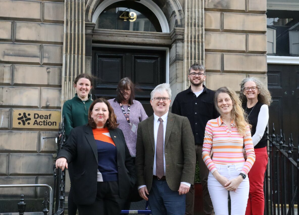 A group of people stand outside a building. On the wall is a plaque that read "Deaf Action".