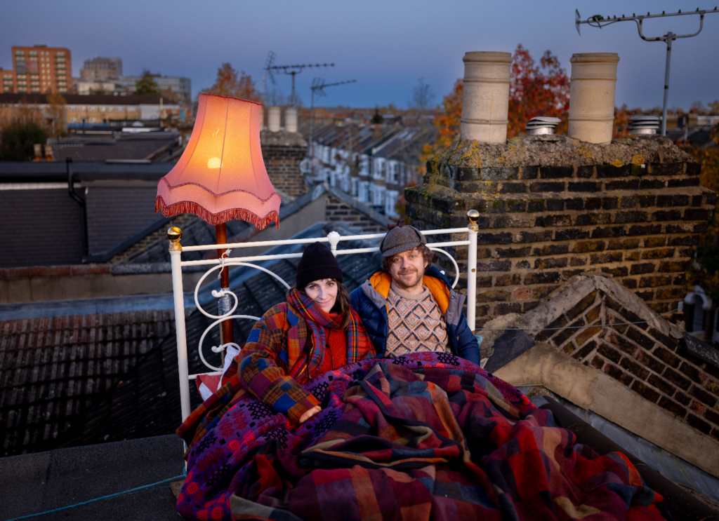 Two people lying in a bed on the roof of a house. Both are wearing winter clothes and hats. There is a lamp shade beside them and in the background are houses and chimney tops.
