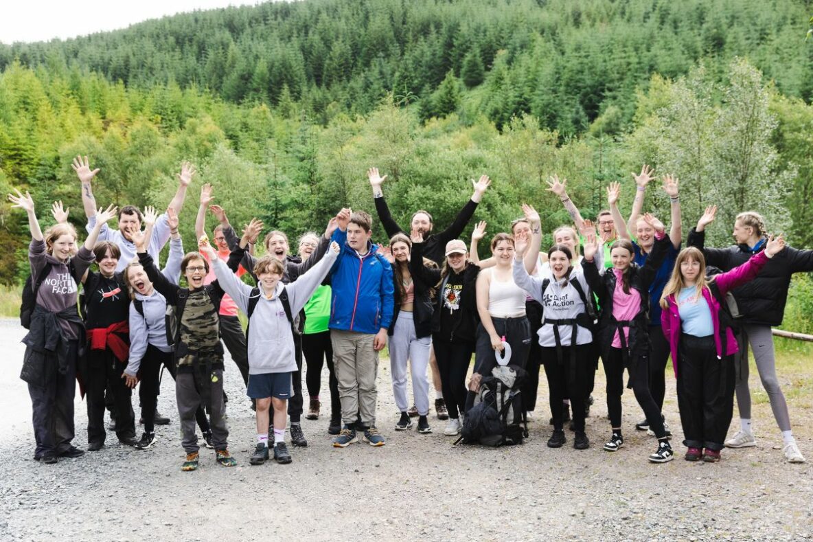 A group of children with their hands in the air. They are standing outside in a landscape of trees and hills. They are wearing outdoor clothes and rucksacks.