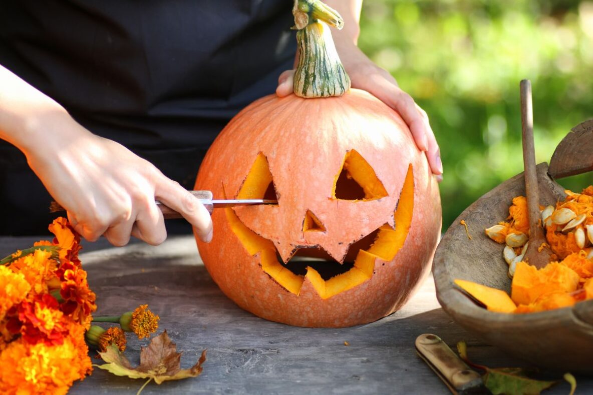 A person carving a pumpkin on a table. Also on the table are orange leaves and a basket.